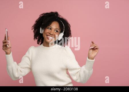 Heureuse femme millénaire à la peau sombre avec casque d'écoute appréciant l'écoute du nouvel album de l'artiste, danse, fait des mouvements à la musique, tenant pho mobile Banque D'Images