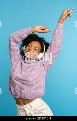 Femme biraciale dynamique qui danse, sourit largement, porte des écouteurs sans fil, a une bonne humeur, garde les mains soulevées dans l'air, isolé sur le backgroun bleu Banque D'Images