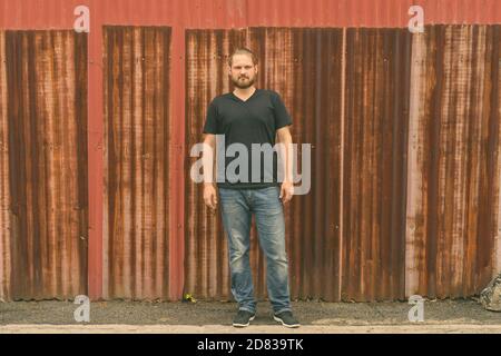 Jeune homme barbu avec cheveux attachés debout contre l'ancien mur feuille rouillée Banque D'Images