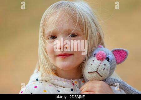 Portrait en gros plan d'une belle petite fille blonde qui embrasse son jouet fait main préféré (chat tricoté) à l'extérieur. Jour venteux, cheveux désordonnés sur le visage. Banque D'Images