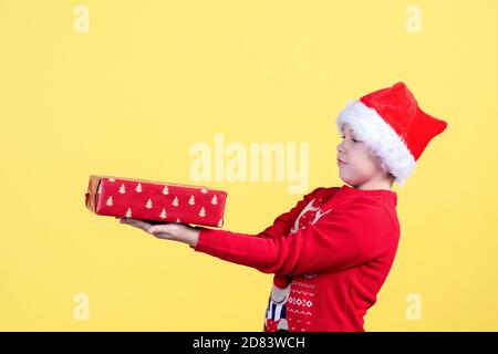 Un enfant émotif surpris dans un chapeau de père Noël tient un coffret cadeau de Noël emballé sur un fond jaune dans le Studio. Copier l'espace. Banque D'Images