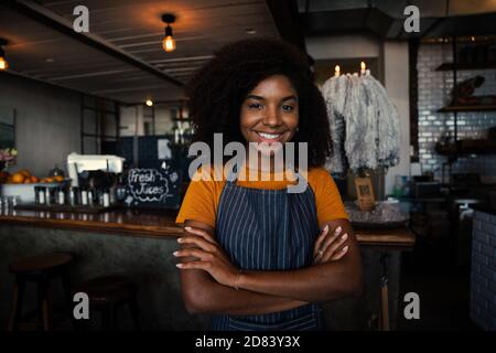 Belle serveuse souriante portant un tablier debout et armé dans un café branché Banque D'Images