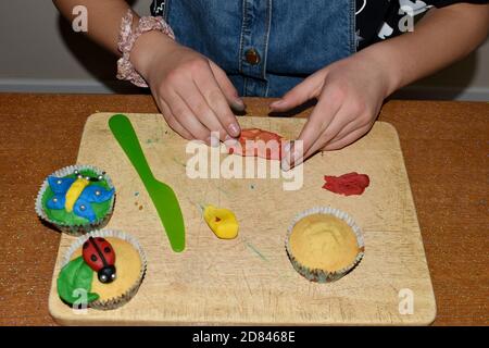 les mains et les bras des enfants font de la garniture givrante pour les petits gâteaux, font de la confection à l'intérieur, avec différentes glaçages aux couleurs vives sur la planche à découper Banque D'Images