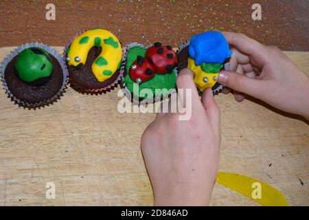 les mains et les bras des enfants font de la garniture givrante pour les petits gâteaux, font de la confection à l'intérieur, avec différentes glaçages aux couleurs vives sur la planche à découper Banque D'Images