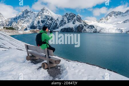 Vue arrière du jeune homme avec veste verte sur le banc au lac Luenersee avec les montagnes des Alpes en arrière-plan pendant la journée de neige. Banque D'Images