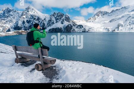 Vue arrière du jeune homme avec veste verte sur le banc au lac Luenersee avec les montagnes des Alpes en arrière-plan pendant la journée de neige. Banque D'Images