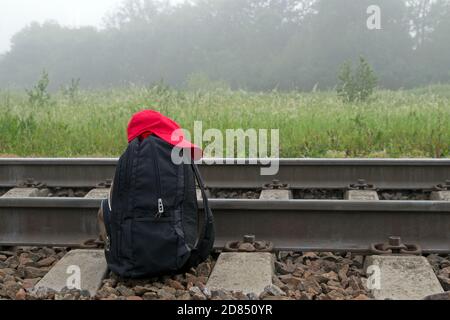 Sac à dos noir avec deux sangles et capuchon rouge sur elle se trouve à côté de voies de chemin de fer et de traîneaux sur un fond de prairie, d'arbres et d'un ciel brumeux. Banque D'Images