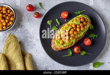 toast à l'avocat avec pois chiches rôtis, herbes, cerise sur une assiette sombre. en-cas végétalien. fond gris, image horizontale Banque D'Images