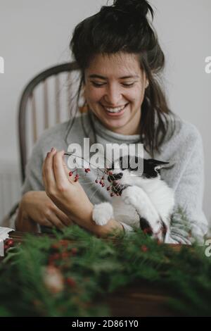 Chat mignon aidant jeune femme heureuse faisant couronne de noël rustique, tenant des baies rouges et des branches vertes, l'avènement des vacances. Image authentique et élégante, pe Banque D'Images