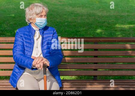 Femme âgée portant un masque médical reposant sur un banc à l'extérieur et tenant un bâton de marche. Femme aux cheveux gris avec masque de protection et canne dans les mains Banque D'Images