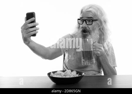 Happy senior man smiling barbu pendant la prise de photo avec téléphone mobile selfies en maintenant le verre de bière avec bol de croustilles de pommes de terre sur la table en bois Banque D'Images