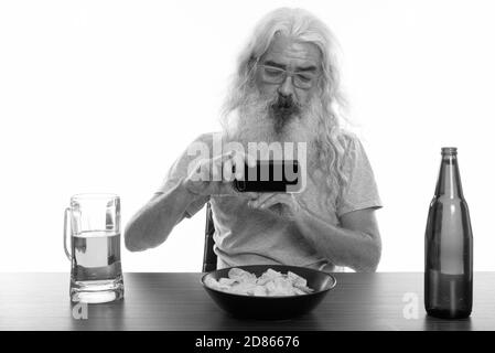 Studio shot of senior homme barbu en photo avec un téléphone mobile de bière et le bol de chips sur la table en bois Banque D'Images