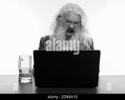 Studio shot of senior homme barbu en colère crier tout en utilisant un ordinateur portable avec verre de bière sur la table en bois Banque D'Images