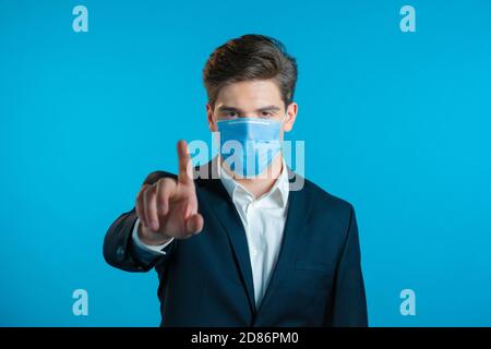 Portrait d'un homme d'affaires sérieux en costume professionnel et masque médical montrant le rejet du geste par le signe d'arrêt du doigt. Homme isolé sur fond bleu. Banque D'Images