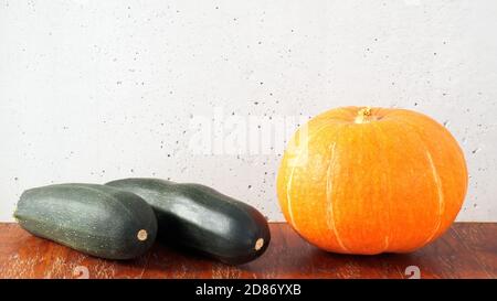 Citrouille orange et deux courgettes vert foncé sur une table en bois sur fond de mur en béton gris Banque D'Images