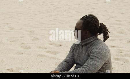 Jeune homme noir afro-américain assis sur le sable en chandail et reposant sur les vacances d'hiver. Photo de haute qualité Banque D'Images