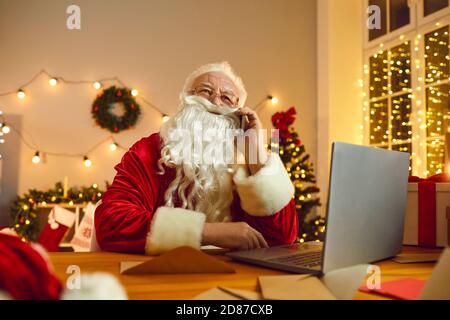 Le Père Noël travaille sur un ordinateur portable tout en parlant au téléphone dans une chambre décorée pour Noël. Banque D'Images