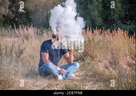 Homme moderne soufflant beaucoup de fumée en utilisant la cigarette électronique de la vape dans la forêt. L'homme aime vraiment le processus de fumer. Banque D'Images