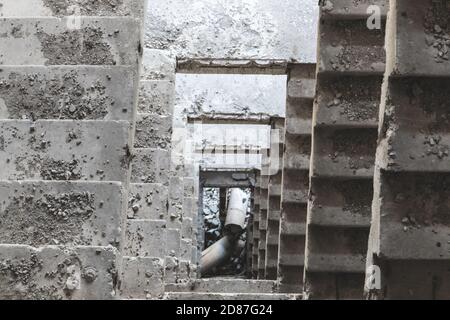 Escalier en béton poussiéreux en niveaux de gris à l'intérieur d'un bâtiment abandonné en ruines. Vue vers le bas tunnel de perspective a traversé la maison de plusieurs étages Banque D'Images