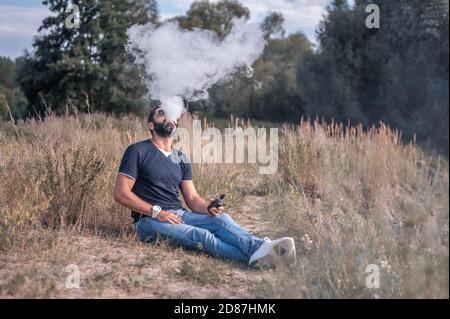 Beau homme de vape appréciant un dispositif électronique de fumée sur l'herbe. Non-fumeurs. Banque D'Images