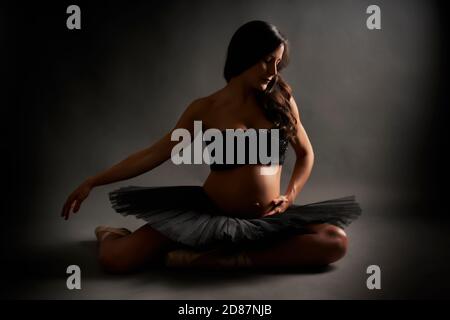 Jeune ballerine enceinte effectuant une posture classique de ballet avec le dessus noir et tutu en studio Banque D'Images