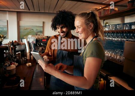 Serveur homme et femme joyeux portant un tablier avec une tablette numérique derrière le comptoir dans le café Banque D'Images