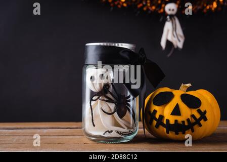Drôle Halloween jour décoration fête, le fantôme blanc visage effrayant et araignée noire en verre pot et potiron dehors côté sur table en bois, studio photo isola Banque D'Images