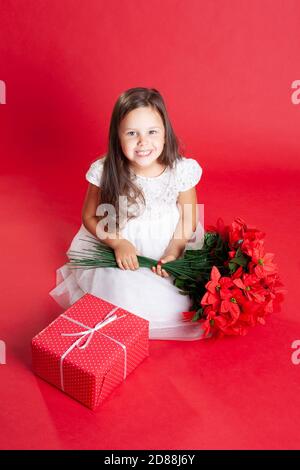 un enfant vêt d'une robe blanche rie et apprécie cadeaux et fleurs sur fond rouge Banque D'Images