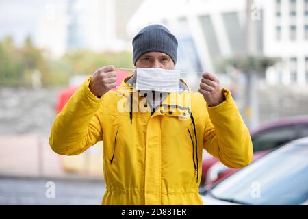 Beau barbu homme regarde la caméra et tient un masque médical devant lui. Arrière-plan flou en extérieur. L'homme dans la rue doute si un masque Banque D'Images