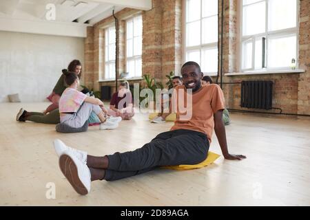 Portrait de l'homme africain souriant à la caméra assis sur le sol en studio de danse avec d'autres personnes en arrière-plan Banque D'Images