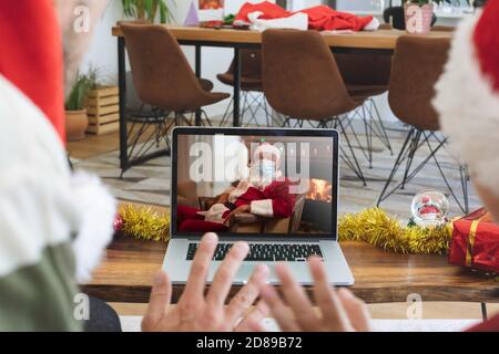 Vue arrière de l'homme et du fils dans les chapeaux de santa Waving tout en ayant un vidéocall avec le père noël portant le visage Banque D'Images