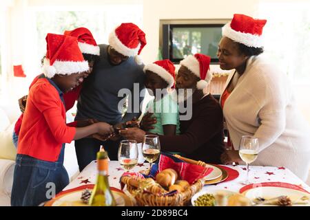 Famille de plusieurs générations portant chapeau de père noël regardant dans le smartphone tout en se tenant près de la table à manger dans le salon à la maison. noël festivit Banque D'Images