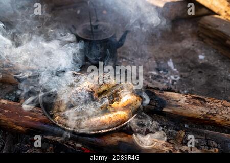 De gros morceaux de poisson fraîchement pêchés sont frits dans une poêle au-dessus d'un feu de camp, à côté d'une bouilloire, dans un camping, un jour d'été. Banque D'Images