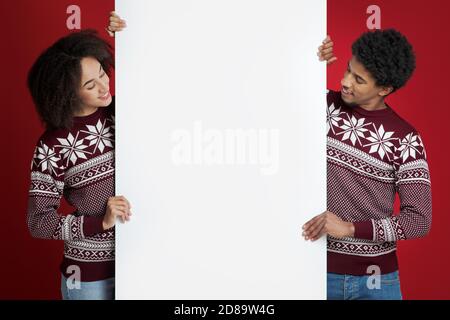 Jeunes couples afro-américains amicaux dans des chandails identiques de Noël tient tableau vierge pour les mots et regarde la bannière Banque D'Images