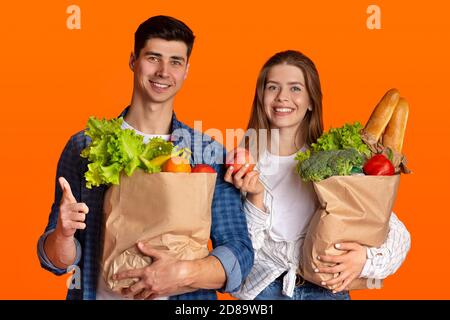 Joyeux couple millénaire amoureux des sacs en papier, dame tenant la pomme, gars pointe son doigt vers l'appareil photo Banque D'Images