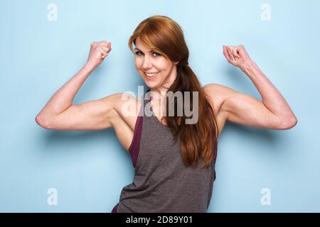 Portrait d'une femme mûre heureuse qui plie les muscles du biceps contre le bleu mur Banque D'Images