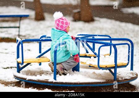 Petite fille triste assise sur un carrousel lors d'une journée d'hiver enneigée. La fille n'est pas heureuse de la première neige Banque D'Images