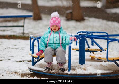 Petite fille triste assise sur un carrousel lors d'une journée d'hiver enneigée. La fille n'est pas heureuse de la première neige Banque D'Images