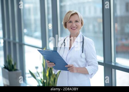 Femme blonde médecin avec stéthoscope debout à côté de la fenêtre, tenant le dossier de clip Banque D'Images