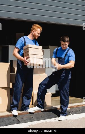 Les déménageurs travaillant avec des boîtes en carton et une presse-papiers à proximité de l'entrepôt à l'extérieur Banque D'Images