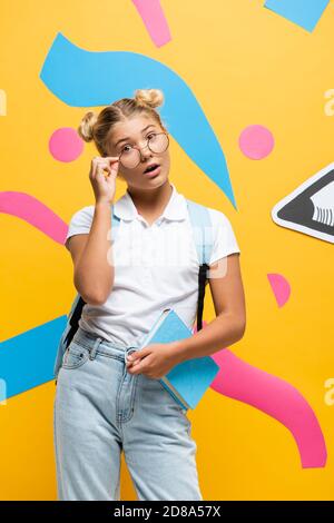 surpris écolière touchant des lunettes et regardant l'appareil photo tout en tenant livre près du crayon de papier et éléments abstraits sur le jaune Banque D'Images