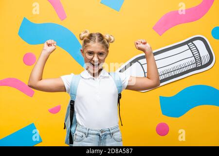 écolière excitée à lunettes montrant le geste du gagnant près du crayon de papier et éléments multicolores sur jaune Banque D'Images