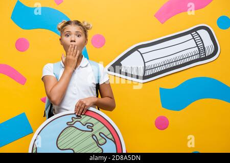 pupille choquée couvrant la bouche avec la main tout en tenant le globe maquette près du crayon de papier et des éléments décoratifs sur le jaune Banque D'Images
