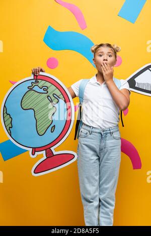 choqué écolière couvrant la bouche avec la main tout en tenant la maquette de globe près du crayon de papier et des éléments décoratifs sur le jaune Banque D'Images