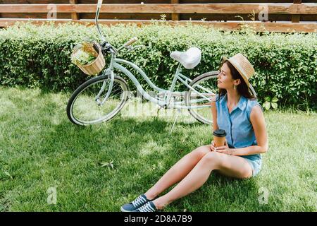 femme en chapeau de paille tenant une tasse en papier et assise sur herbe près de la bicyclette Banque D'Images