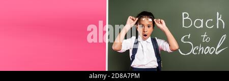 choqué afro-américain écolière ajuster des lunettes près de l'inscription à l'école sur tableau noir vert sur fond rose, photo panoramique Banque D'Images