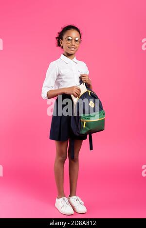 souriante écolière afro-américaine en verres mettant un livre dans un sac à dos sur rose Banque D'Images