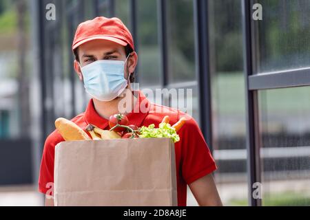 Coursier dans un masque médical contenant un sac à provisions avec des légumes frais sur la rue urbaine Banque D'Images