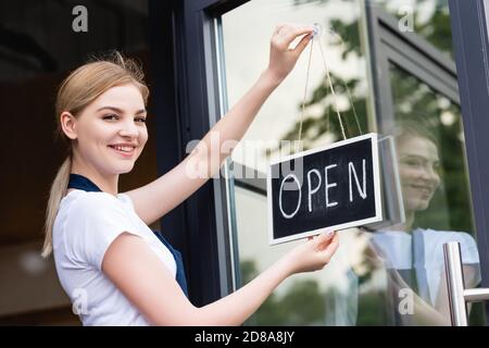 Vue latérale de la serveuse souriant à l'appareil photo tout en accrochant une pancarte avec inscription ouverte sur la porte du café Banque D'Images