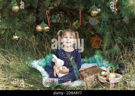 Jolie petite fille assise dans l'herbe près de l'arbre de Noël et des jouets et des cadeaux et heureux. Décoration arbre de Noël. Enfant souriant. joyeux noël. Enfant Banque D'Images